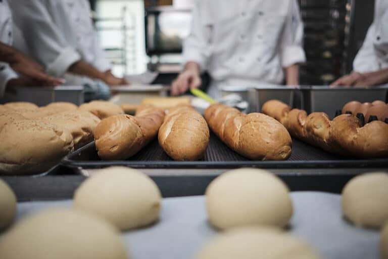 Close-up shot and selective focus at baguettes and delicious bread in front of white uniform chefs team, kneading raw pastry dough, preparing fresh bakery food, baking in oven in restaurant kitchen.
